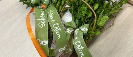 Zwei Ostersträuße mit grünen Bändern, einer mit der Aufschrift 'Schone Ostern' und der andere mit 'Frohe Ostern', stehen auf einem Tisch mit einem weißen Teller und Messer.