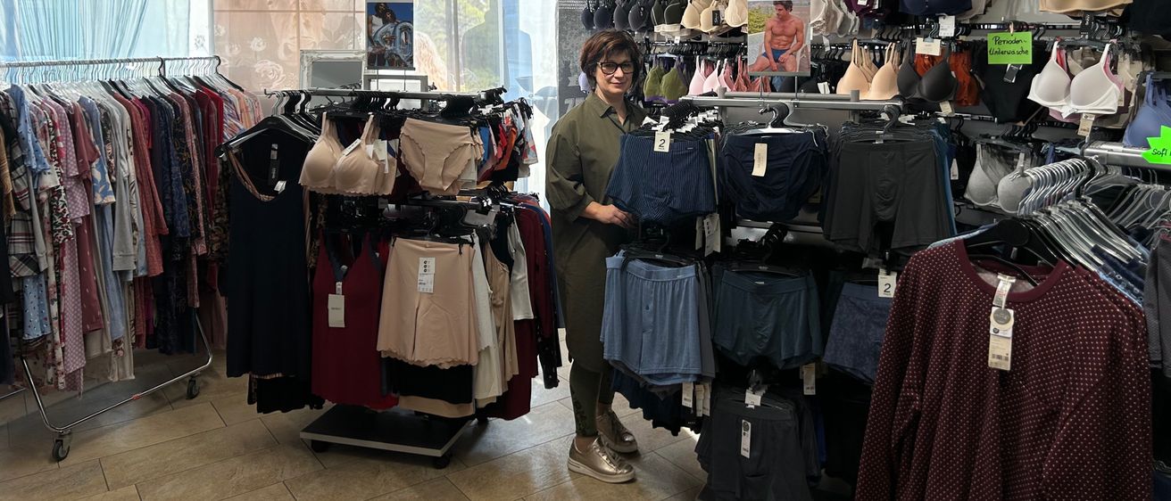 Eine Frau steht in einem Bekleidungsgeschäft mit verschiedenen Kleidungsstücken zur Schau, darunter Dessous, Röcke und Blusen. Der Laden hat ein modernes Design mit weißen Wänden und gefliesten Böden.