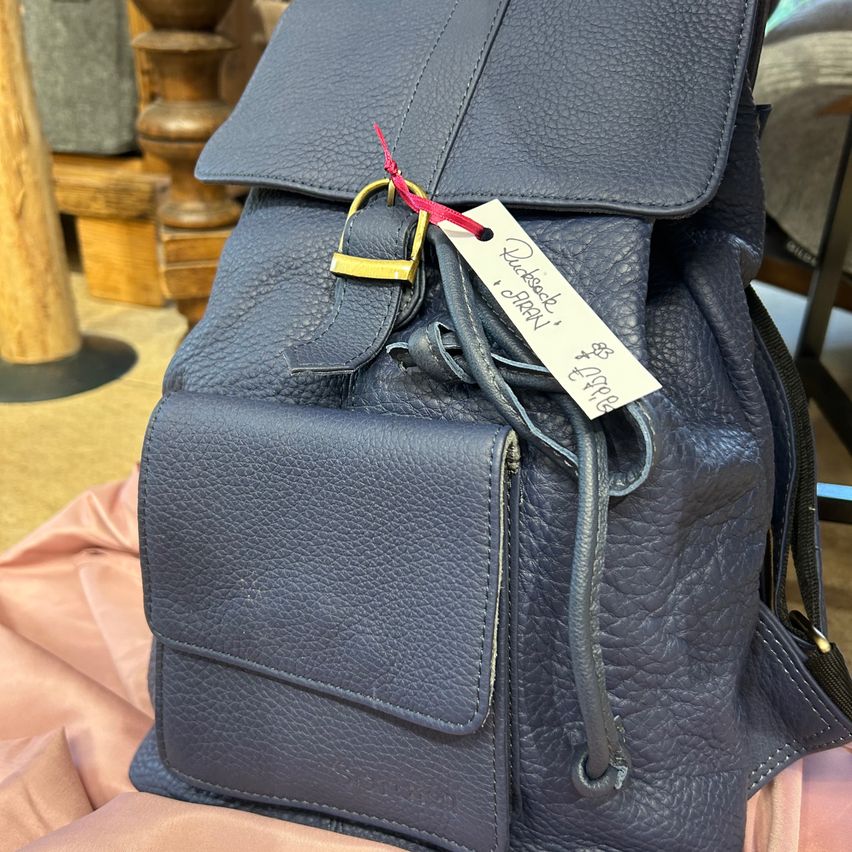 A black leather backpack with a gold buckle and a white tag reading 'Quidsecki' by 'Orion' is displayed on a pink surface.