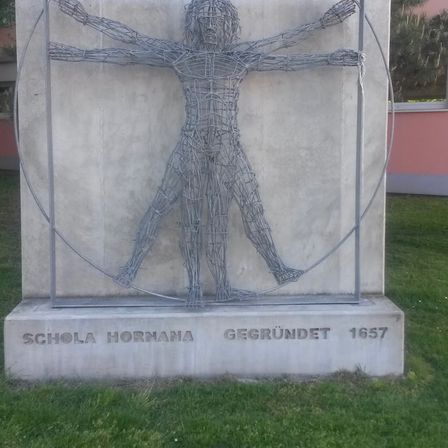Eine Skulptur einer menschlichen Figur aus Draht, eingebettet in einen Betonrahmen. Darunter steht 'SCHOLA HORNANA GEGRUNDET 1657'.