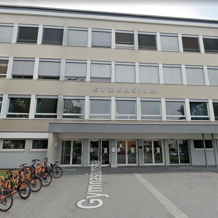 Ein großes Gebäude mit dem Wort Gymnasium an der Fassade, mehrere Fahrräder vor dem Gebäude und verglaste Türen.