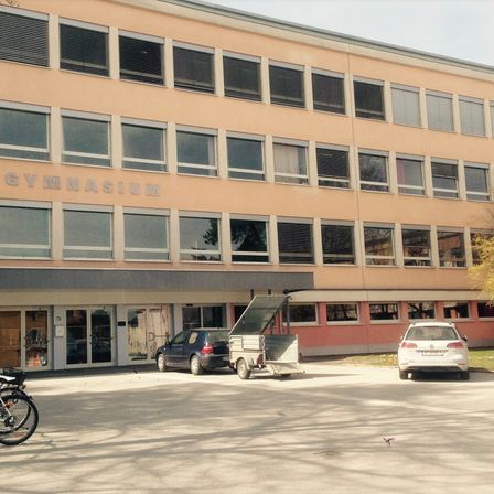 Ein Gebäude mit dem Wort Gymnasium darauf. Vor dem Gebäude sind zwei Autos geparkt, und ein Anhänger ist an eines von ihnen angeschlossen. Ein Fahrrad steht links.