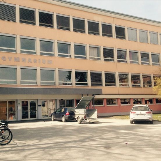 Ein Gebäude mit dem Wort Gymnasium darauf. Vor dem Gebäude sind zwei Autos geparkt, und ein Anhänger ist an eines von ihnen angeschlossen. Ein Fahrrad steht links.