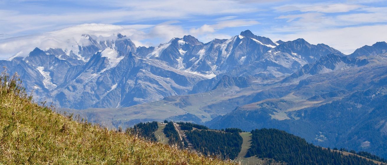 Ein Panoramablick auf eine bergige Landschaft unter einem blauen Himmel mit verstreuten Wolken. Schneebedeckte Gipfel und üppige Grünflächen dominieren die Szene.
