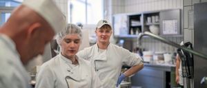 Zwei Köche in einer Küche, einer mit Haarnetz und der andere mit Mütze, lächeln. Hinter ihnen ist ein Waschbecken und ein Fenster zu sehen.