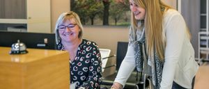 Zwei Frauen sind in einem Büro, eine arbeitet an einem Laptop, die andere lächelt und berührt den Laptop auf dem Schreibtisch. Hinter ihnen ist eine Wand mit einem großen Gemälde eines Baumes.
