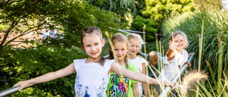 Mehrere junge Mädchen stehen lächelnd in einem Garten. Ein Mädchen mit ausgestreckten Armen winkt. Der Garten ist üppig mit grünen Bäumen und Pflanzen.