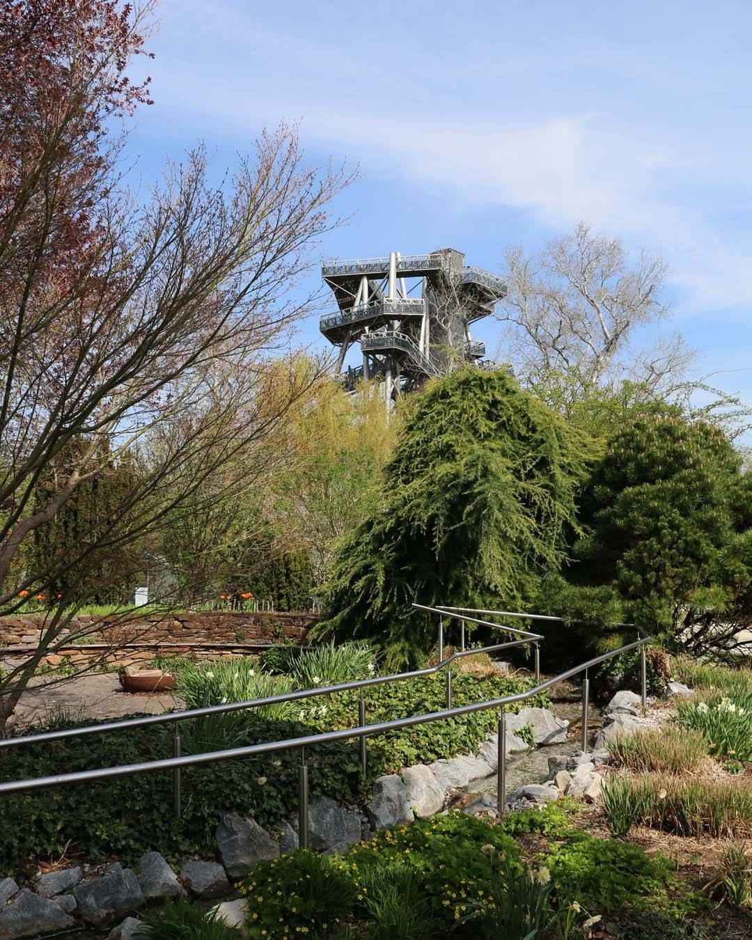 Ein hoher Turm ist von Bäumen und Pflanzen umgeben, mit einem kleinen Bach vor ihm.