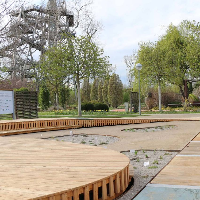 Ein hölzerner kreisförmiger Weg in einem Park, umgeben von Bäumen, Bänken und einem Turm mit Treppen.