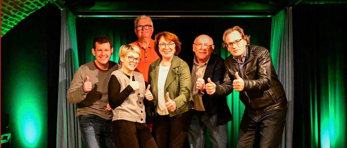 Sechs Personen stehen in einer Reihe, lächeln und geben Daumen hoch, vor einem grünen Hintergrund mit Vorhängen.