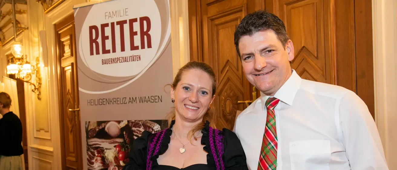 Ein Mann und eine Frau stehen nebeneinander vor einer Holztür und halten eine Wurstgirlande. Die Frau trägt ein traditionelles schwarzes Kleid mit einem lila Saum, und der Mann ist in einem weißen Hemd mit einer rot-grünen Krawatte. Hinter ihnen steht ein Schild mit dem Wort Reiter.