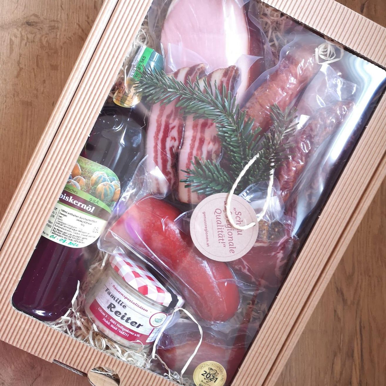 Eine Holzkiste enthält verschiedene Fleischsorten, darunter Speck und Würste, mit einem Glas Marmelade, einer Flasche Saft und einem dekorativen Tannenzweig.