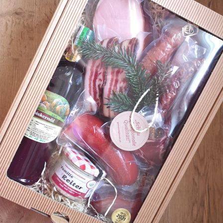 Eine Holzkiste enthält verschiedene Fleischsorten, darunter Speck und Würste, mit einem Glas Marmelade, einer Flasche Saft und einem dekorativen Tannenzweig.