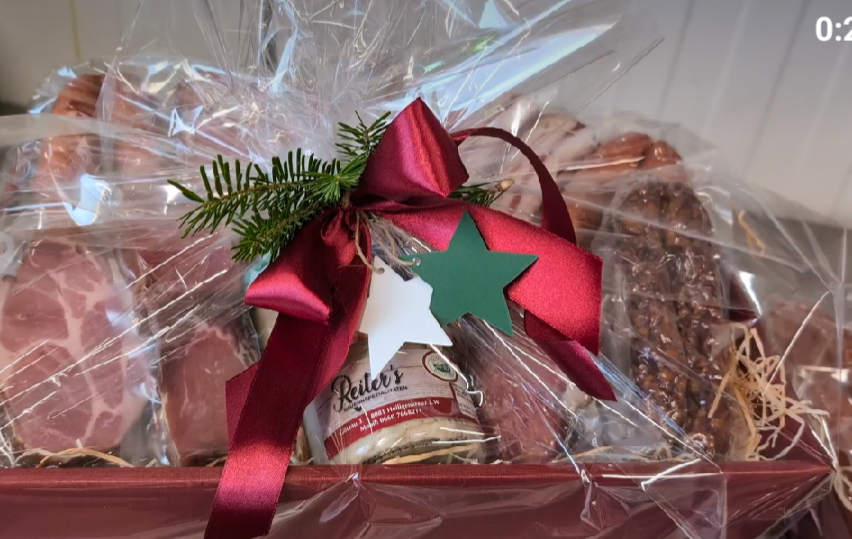 A red gift basket wrapped in clear plastic includes various meats, a jar of Reiler's honey mustard, and a festive green star on a red ribbon.