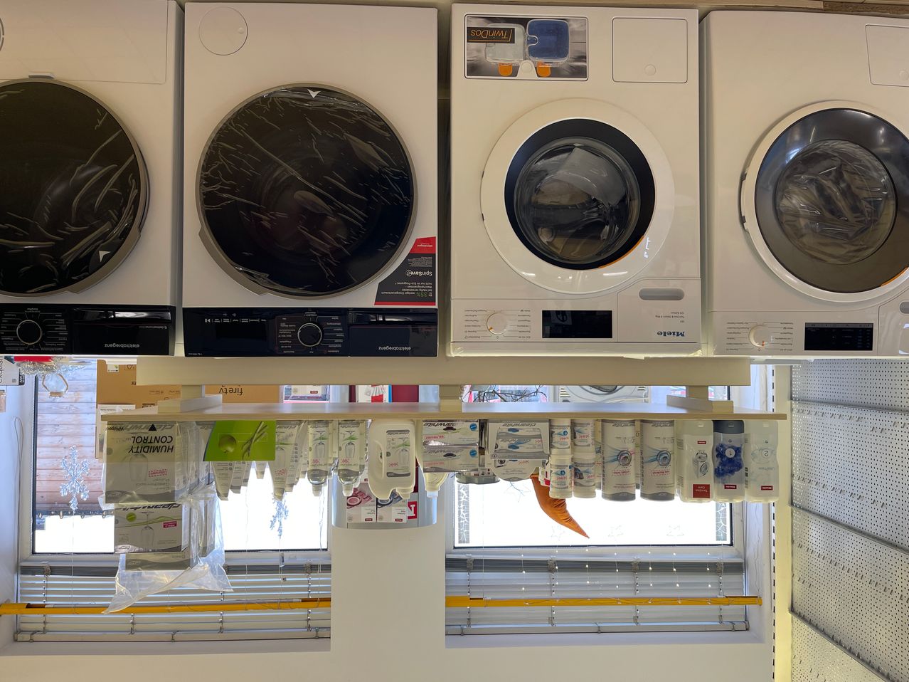 Vier Waschmaschinen sind in einer Reihe aufgestellt. Die erste hat eine schwarze Abdeckung, und die zweite hat eine weiße Abdeckung. Beide sind neu und haben Etiketten auf der Vorderseite. Unter den Maschinen sind verschiedene Waschmittel und andere Produkte ausgestellt.