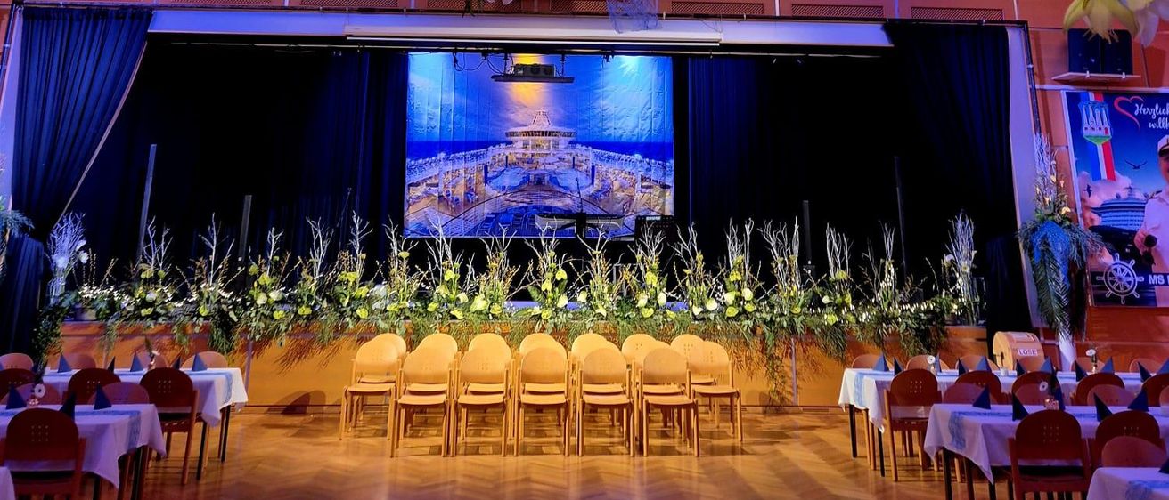 Ein Bankettsaal mit Tischen, Stühlen, Blumen und einer Leinwand, die eine Meereslandschaft zeigt. Die Decke ist mit hängenden Blumen und Lichtern geschmückt.