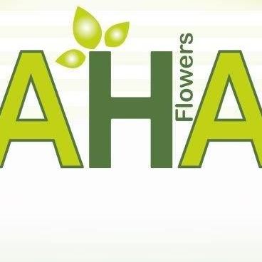 Das Bild zeigt das Logo von AHA Flowers, das einen stilisierten grünen Buchstaben 'A' mit einem Blatt darüber darstellt. Die Buchstaben sind fett und grün auf weißem Hintergrund. Der Text 'Flowers' steht in Grün auf der rechten Seite des Buchstabens 'A'.