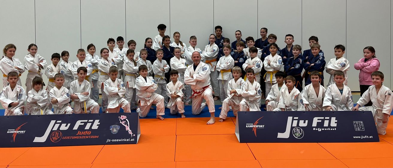 Eine Kampfsportklasse posiert für ein Foto. Erwachsene und Kinder in Uniform stehen und hocken auf einem orangefarbenen Teppich. Ein Mann in der Mitte mit verschränkten Armen.