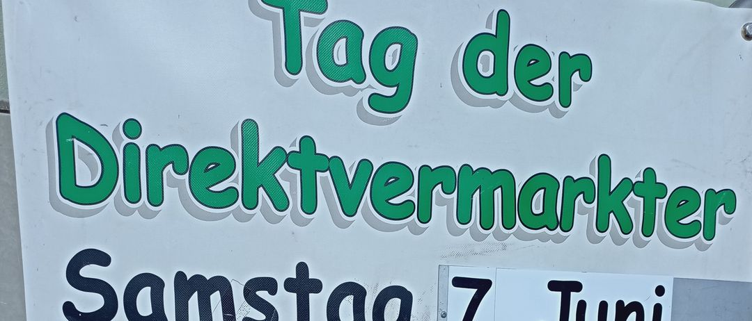 Ein Schild bewirbt einen Direktmarkt am Samstag, 7. Juni, von 9 Uhr bis 13 Uhr. Es listet die Adresse von Familie Menitz als Eisenstädterstr. 18, 2485 Wimpassing auf. Das Schild zeigt verschiedene Lebensmittel wie Brot, Fleisch, Würste und Gläser mit Marmeladen sowie Blumen und Wein.