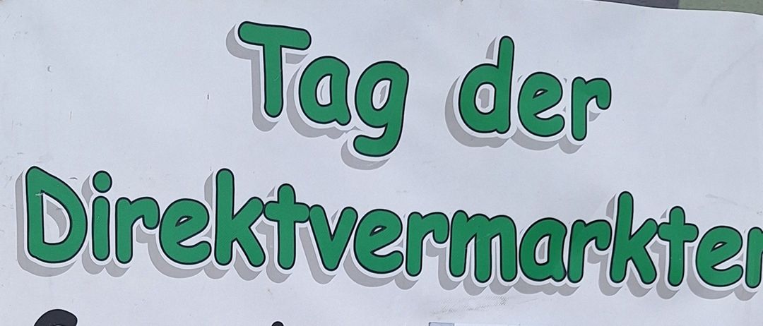 Ein Schild bewirbt einen Direktmarkt am Samstag, 11. Oktober, von 9 Uhr bis 13 Uhr. Es zeigt eine Familie namens Menitz, ansässig in Eisenstadterstr. 18, 2485 Wimpassing. Der Plakat zeigt verschiedene Lebensmittel wie Fleisch, Würste, Marmeladen und Beeren.