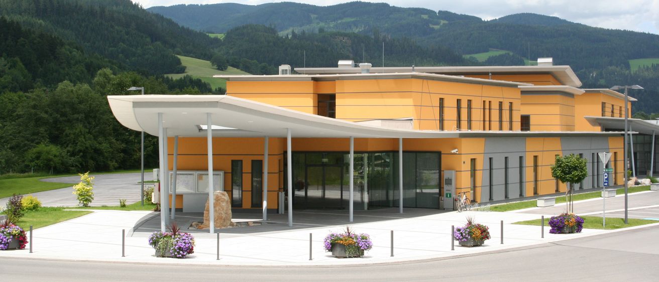 Ein modernes gelbes Gebäude mit Glastüren und Fenstern steht in einem Gebirgsgebiet. Davor befindet sich ein überdachter Eingang mit Topfpflanzen und einer Steinskulptur.