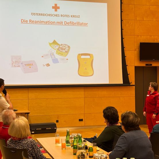 Eine Präsentationsfolie mit dem Text 'Die Reanimation mit Defibrillator' wird auf einem Bildschirm angezeigt. Vor dem Bildschirm sitzen mehrere Erwachsene auf Stühlen.