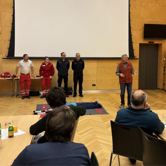 Eine Gruppe von Menschen ist in einem Raum versammelt. Ein Mann steht und spricht, während andere auf Stühlen sitzen. Eine Person liegt auf dem Boden.