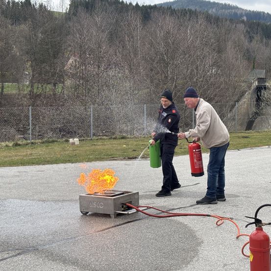 Zwei Personen stehen neben einem Feuerlöscher. Einer von ihnen spritzt Wasser auf das Feuer, während der andere einen Feuerlöscher in der Hand hält.