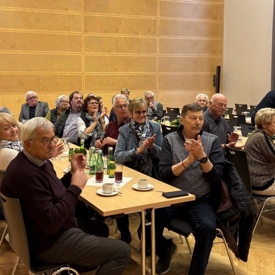 Eine Gruppe älterer Menschen sitzt um einen Tisch mit Tassen, Untertassen und Flaschen und klatscht und lächelt möglicherweise.