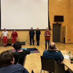 Fünf Männer in roten und schwarzen Anzügen stehen vor einer Beamerleinwand. Ein Mann liegt auf einer Matte auf dem Boden. Vor den Männern sitzen mehrere Personen in Stühlen.