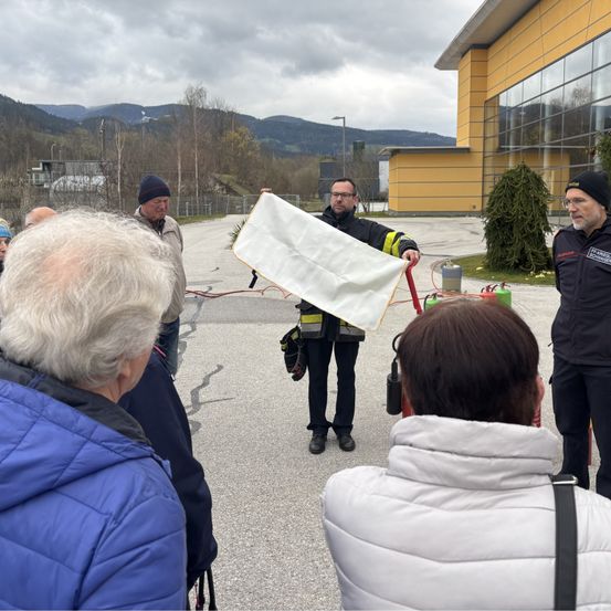 Ein Feuerwehrmann hält ein weißes Tuch und erklärt etwas einer Gruppe von Menschen auf einem Parkplatz.