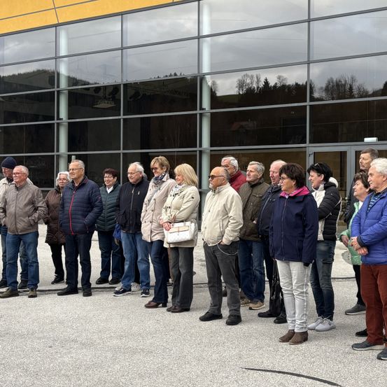 Eine Gruppe Erwachsener steht vor einem Gebäude mit Glaswänden. Das Gebäude hat ein modernes Design mit einem Gelb- und Grautonschema.