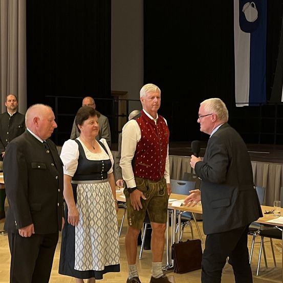 Ein Mann mit einem Mikrofon spricht zu einer Frau in traditioneller deutscher Kleidung, einem Mann in einer roten Weste und einem anderen Mann in einem Anzug. Dahinter steht ein Tisch mit einem Glas und einer Tasche.