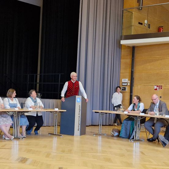 Ein Mann steht hinter einem Podium mit einem Mikrofon vor einer Gruppe von Personen. Sie sitzen um einen Tisch herum und scheinen eine Diskussion zu führen.