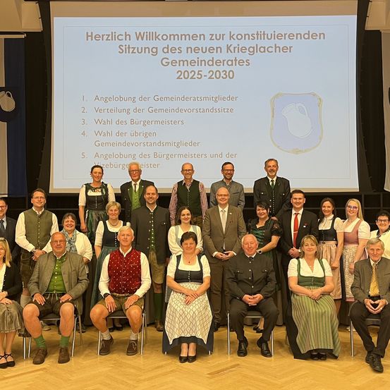 Gruppenfoto von Beamten und Gemeinderatsmitgliedern in traditioneller Kleidung. Im Hintergrund zeigt eine Projektionsleinwand die Tagesordnung für die Sitzung.