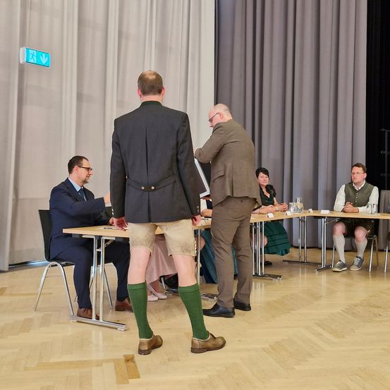 Eine Gruppe von Männern und Frauen sitzt an einem Tisch in einem Raum. Der Mann, der mit dem Rücken zur Kamera steht, trägt Shorts und grüne Socken.