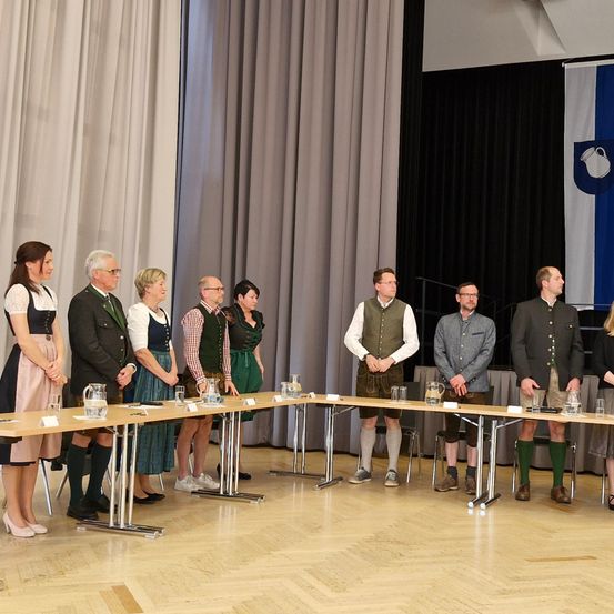 Eine Gruppe Erwachsener in traditioneller bayerischer Kleidung, die hinter einem Tisch mit Gläsern und Krügen in einem Raum mit Holzboden steht.