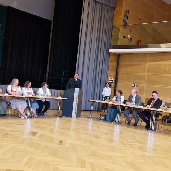 Eine Konferenz findet in einem Saal mit Holzböden statt, wo ein Mann an einem Podium eine Gruppe von Personen an Tischen anspricht.