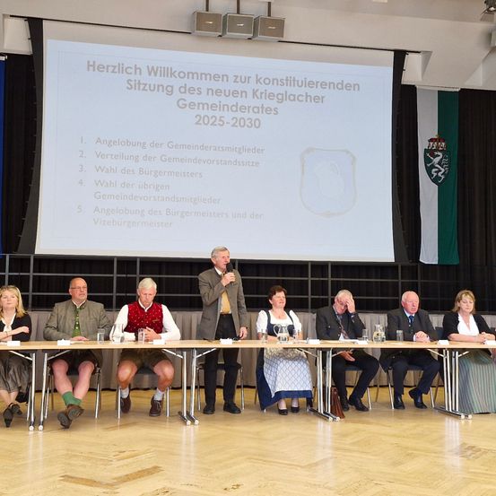 Eine Veranstaltung mit Menschen in traditioneller Kleidung, die an einem Tisch sitzen, während ein Mann mit Mikrofon vorne steht, neben einer Leinwand mit den Worten 'Herzlich Willkommen'.