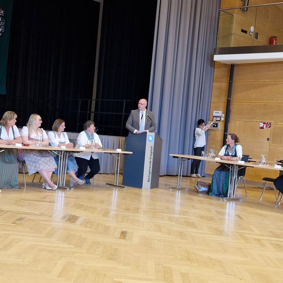 Eine Gruppe von Personen sitzt an Tischen, während ein Mann an einem Podium spricht. Dahinter befindet sich ein Vorhang und ein Geländer, und eine Frau steht hinter der Gruppe.