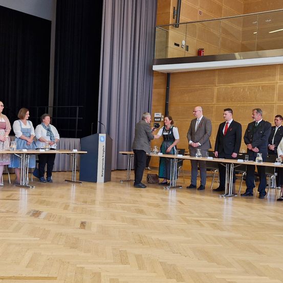 Eine Gruppe von Menschen in Anzügen und Kleidern steht hinter einem Tisch in einem Konferenzsaal, ein Mann hält ein Mikrofon.