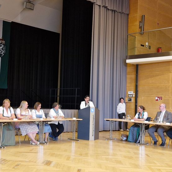 Eine Gruppe von Menschen sitzt um einen Tisch in einem Konferenzsaal. Eine Frau an der Rednertribüne spricht in ein Mikrofon. Hinter der Tribüne hängt ein Banner an der Wand.