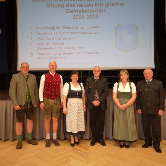 Eine Gruppe von Menschen in traditioneller Kleidung steht vor einer Projektionsleinwand mit einer Liste von Tagesordnungspunkten für den neuen Krieglacher Gemeinderates für 2025-2030.