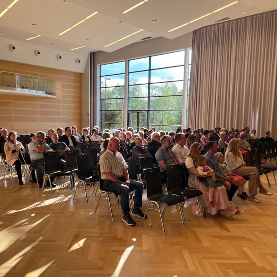 Ein Publikum sitzt in einem großen Saal mit Holzboden, Glasfenstern und Vorhängen. Sie lauschen einem Redner, und einige tragen Brillen, Turnschuhe und Mützen.