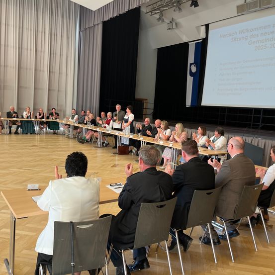 Eine Gruppe von Menschen versammelt sich in einem Besprechungsraum, einige klatschen und andere sitzen in Stühlen. Eine Projektionsleinwand an der Wand zeigt Text.