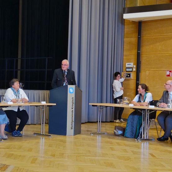 Ein älterer Mann steht an einem Rednerpult und spricht zu einem Gremium von vier Personen, die an einem Tisch sitzen. Der Raum ist mit einer Bühne, Vorhängen und einem Holzboden ausgestattet.