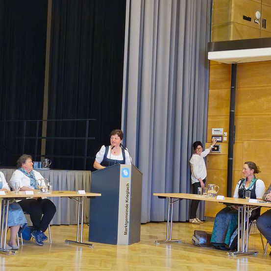 Eine Frau spricht an einem Podium vor einer Gruppe von Frauen, die an einem Tisch sitzen.