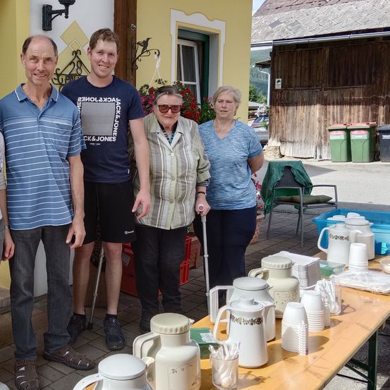 Fünf Personen stehen vor einem Haus mit gelber Wand. Ein Tisch davor ist mit Tassen, Tellern und Teekannen besetzt. Eine Frau benutzt einen Stock. Hinter ihnen ist ein Auto geparkt, und es gibt Mülleimer.