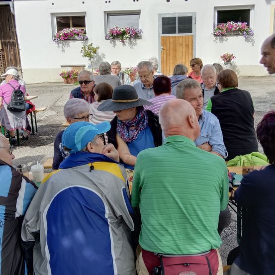Eine Gruppe von Menschen versammelt sich in einem Restaurant und sitzt um Tische herum. Einige tragen Brillen und Hüte. Eine Frau in einem blauen Kleid trägt eine Tasche.