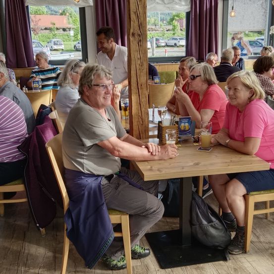 Eine Gruppe von Menschen sitzt an einem Holztisch in einem Restaurant. Ein Mann und eine Frau lächeln und schauen sich an. Biergläser stehen auf dem Tisch.